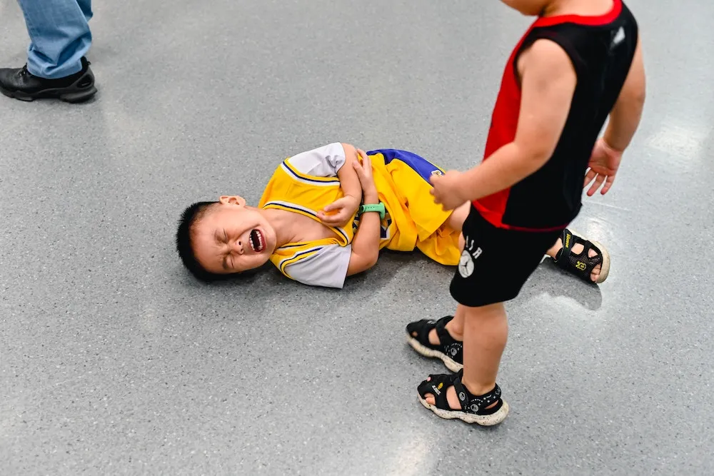 A screaming kid on the floor holding his tummy while another kid stands over him.